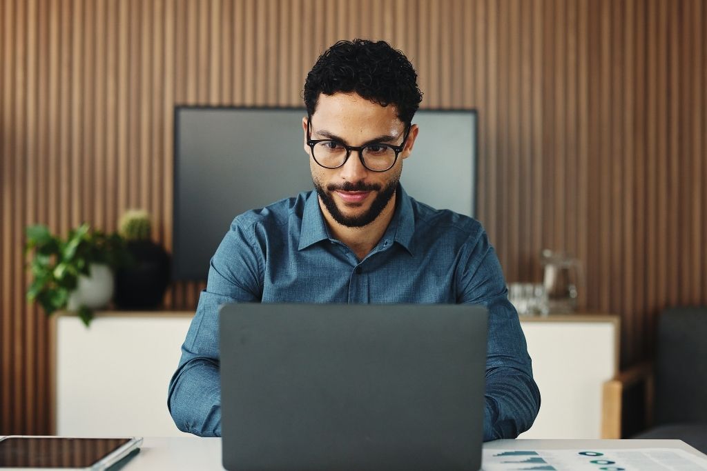 Um homem jovem com óculos e camisa azul trabalha concentrado em seu laptop em um ambiente de escritório moderno. A cena representa a praticidade de gerenciar uma carteira de fundos imobiliários de forma digital, comparando a facilidade do investimento financeiro com a gestão tradicional de imóveis físicos.