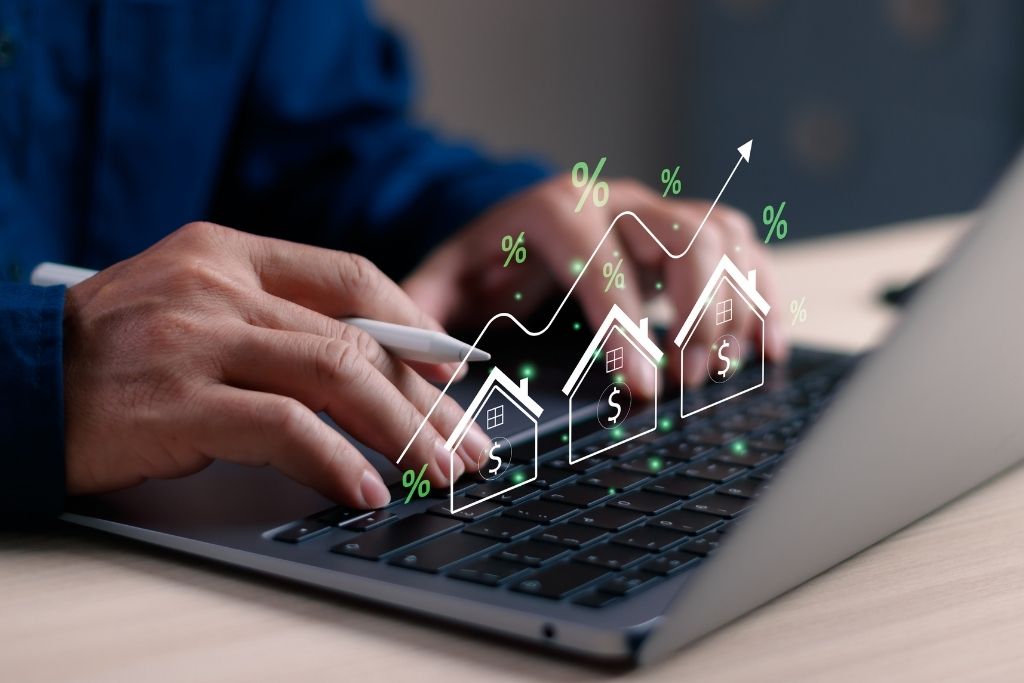 Close nas mãos de uma pessoa utilizando um notebook para análise financeira. Sobre o teclado, elementos digitais flutuantes exibem casas com o símbolo de cifrão, sinais de porcentagem e uma linha de tendência ascendente. A imagem ilustra o processo de avaliação de rentabilidade e indicadores comuns aos diferentes tipos de fundos imobiliários durante o monitoramento de uma carteira de investimentos.