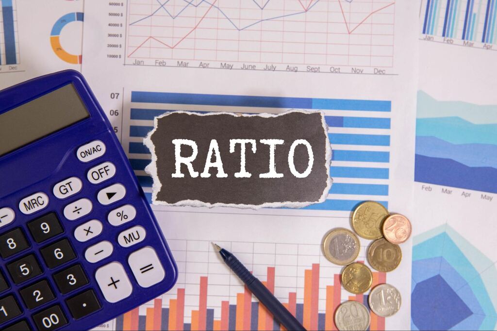 Mesa de escritório com moedas, calculadora e papeis. Na frente, um papel com a palavra "Ratio" escrita em fundo preto.