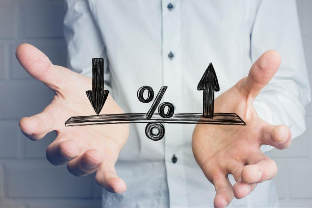 Detalhe de mãos masculinas com as palmas para cima, e um desenho digital de uma gangorra com setas para cima e para baixo, e um "%" ao meio, representando como investir em juros futuros.