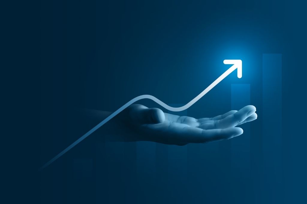 Uma imagem minimalista em tons de azul profundo que representa as oportunidades de investimentos em um cenário de bull market (mercado em alta). Uma mão aberta e iluminada sustenta uma linha gráfica branca que sobe de forma constante e termina em uma seta apontada para cima. Ao fundo, barras verticais semitransparentes reforçam a ideia de progressão financeira e valorização de ativos. A iluminação suave foca no ponto de crescimento, transmitindo uma sensação de otimismo e segurança financeira.