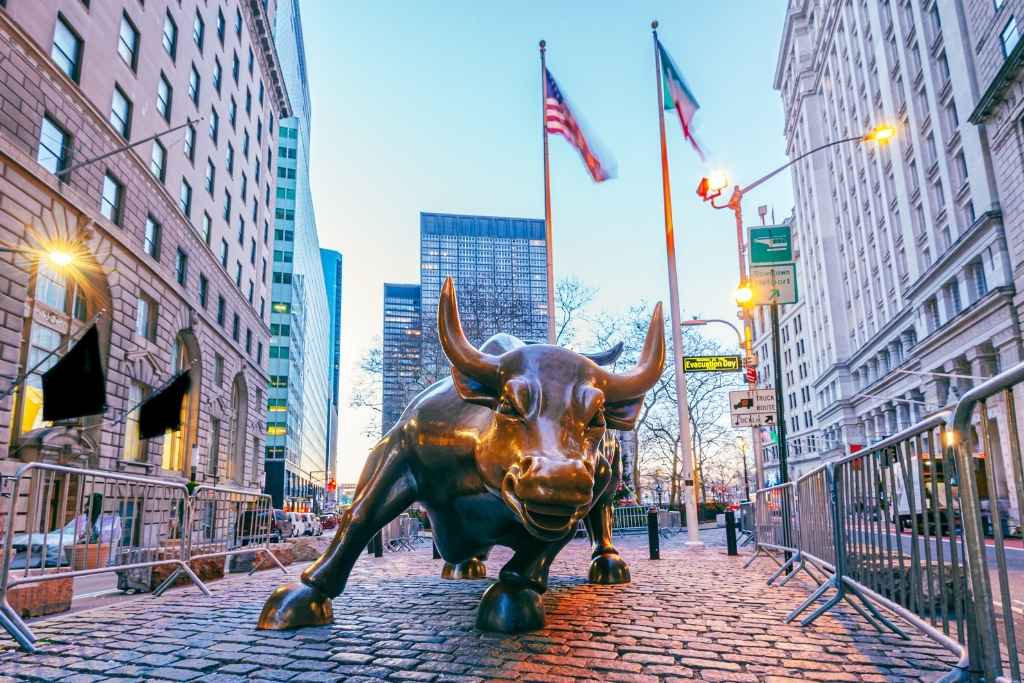 Fotografia da famosa estátua de bronze Charging Bull localizada no distrito financeiro de Nova York. O touro é retratado de frente em uma rua de paralelepípedos entre prédios altos e bandeiras americanas, servindo como o maior ícone mundial do Bull Market e da força da economia global.