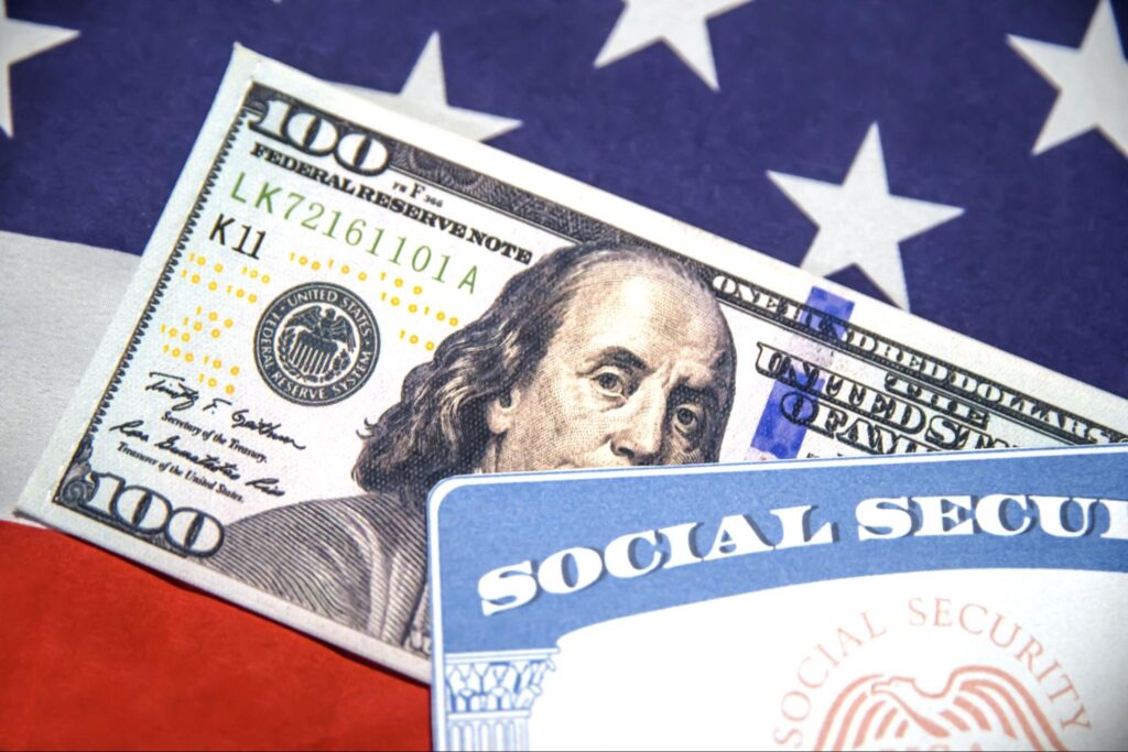 Detalhe de nota de 100 dólares e um cartão de segurança social por cima da bandeira dos Estados Unidos, representando o payroll.