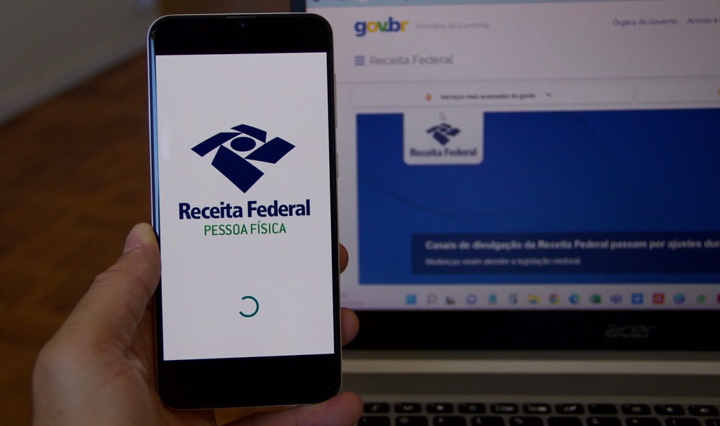 Pessoa segurando celular com logo da Receita Federal na tela, e site da Receita Federal em tela de computador ao fundo.