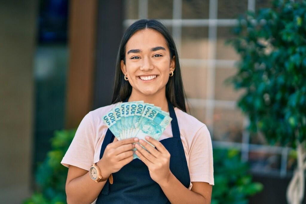 Jovem mulher sorridente, segurando notas de R$ 100, representando onde investir 20 mil reais.