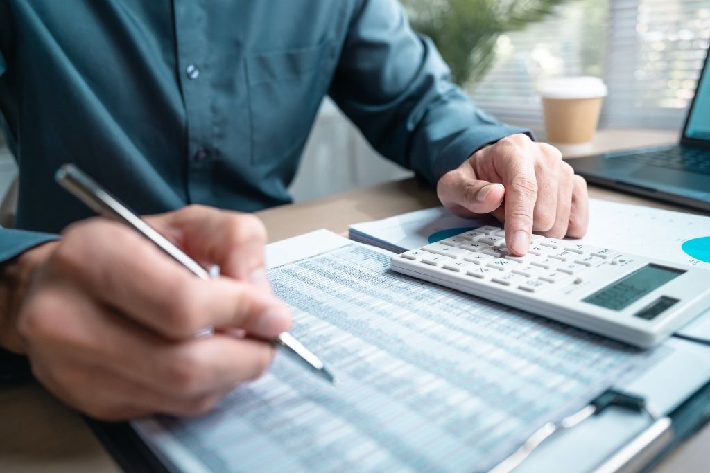 Close focado nas mãos de um profissional que utiliza uma calculadora financeira branca e uma caneta para conferir dados em uma planilha impressa. A imagem detalha o momento técnico de análise de custos e rendimentos, sendo ideal para representar o cálculo do IOF no CDB sobre o lucro de resgates feitos em curto prazo. Ao fundo, um ambiente de escritório iluminado com um notebook e um copo de café reforça o contexto de planejamento financeiro.