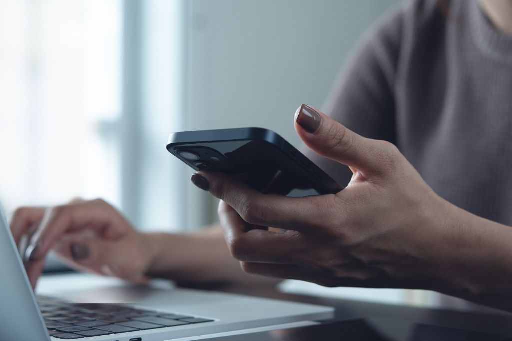 Uma mulher aparece em um plano fechado enquanto utiliza simultaneamente um smartphone e um notebook em sua mesa de trabalho. A composição sugere o acompanhamento de prazos de investimentos em tempo real, focando na estratégia de aguardar o período de isenção do IOF no CDB que ocorre após trinta dias. A iluminação é suave e vem de uma janela lateral, criando um aspecto profissional e sereno.
