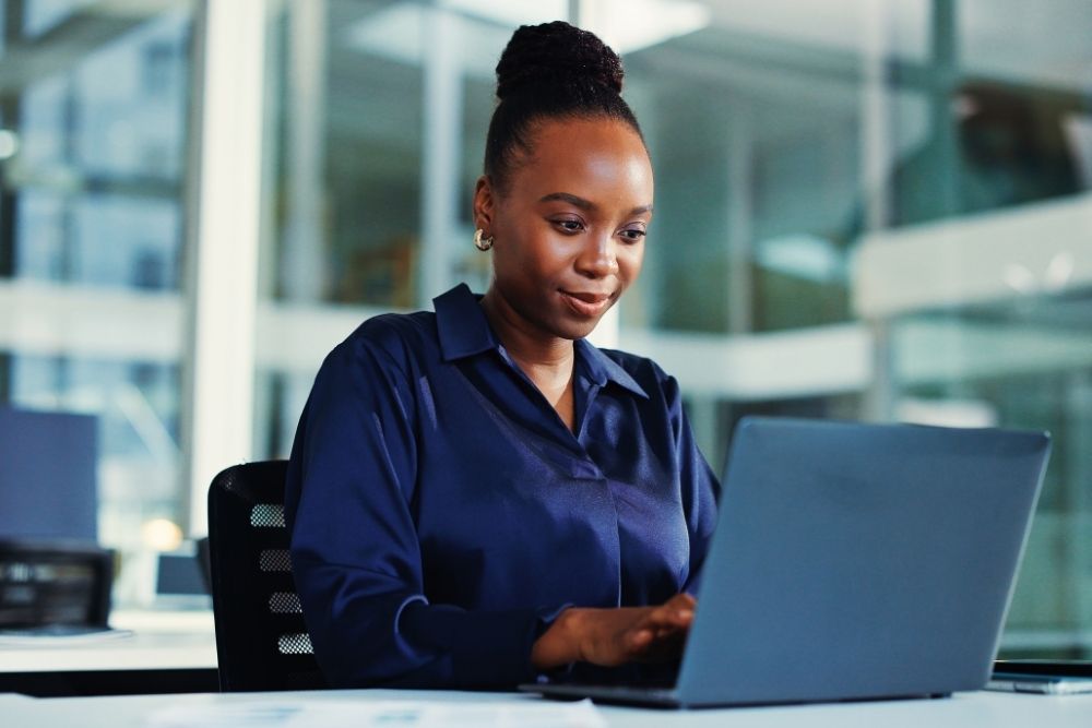 Uma mulher negra com os cabelos presos em um coque sorri levemente enquanto trabalha concentrada em seu notebook em um escritório moderno e iluminado. A cena transmite tranquilidade e autonomia, sugerindo o momento de pesquisa e planejamento financeiro de uma investidora decidindo onde investir 30 mil para alcançar seus objetivos pessoais. O ambiente profissional reforça a seriedade na escolha das melhores opções do mercado.